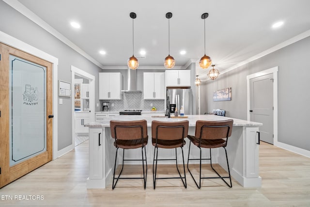 kitchen featuring white cabinetry, stainless steel refrigerator with ice dispenser, a kitchen island with sink, hanging light fixtures, and wall chimney exhaust hood