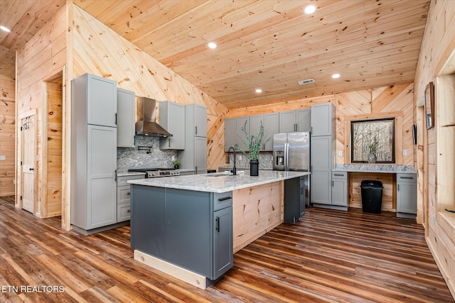 kitchen featuring wall chimney range hood, a center island with sink, wood ceiling, stainless steel appliances, and light stone counters