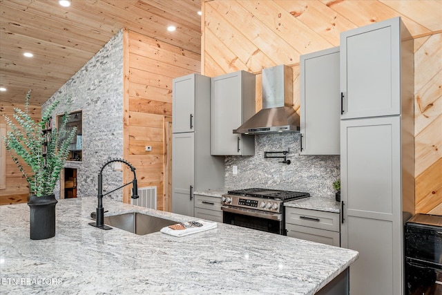 kitchen featuring wood walls, stainless steel gas range, lofted ceiling, wall chimney range hood, and light stone counters