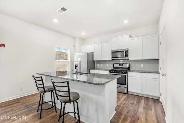 kitchen featuring appliances with stainless steel finishes, a kitchen island with sink, white cabinets, dark stone countertops, and dark hardwood / wood-style floors