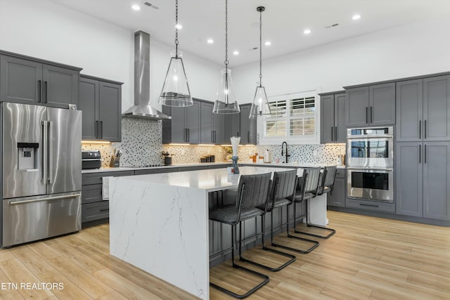 kitchen with hanging light fixtures, a center island with sink, stainless steel appliances, and wall chimney exhaust hood