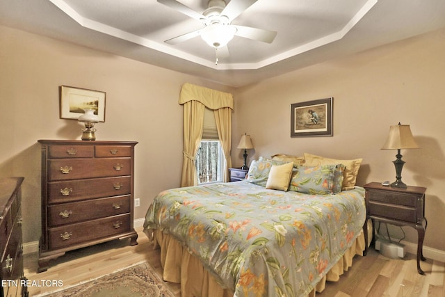 bedroom with light wood-type flooring, a tray ceiling, baseboards, and a ceiling fan