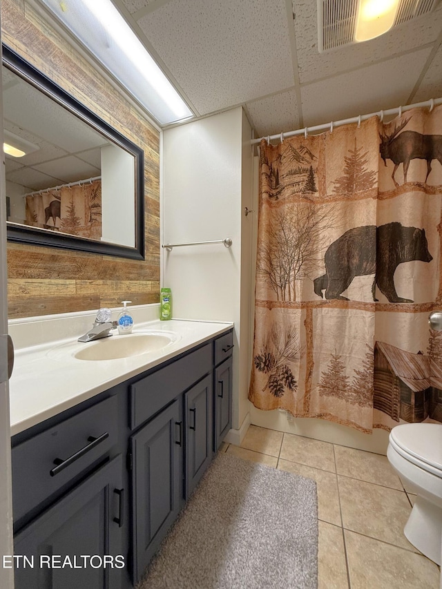 bathroom with toilet, a paneled ceiling, vanity, wooden walls, and tile patterned flooring