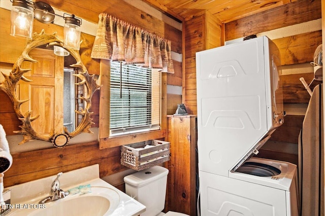 bathroom with stacked washer / drying machine, wooden walls, sink, and toilet