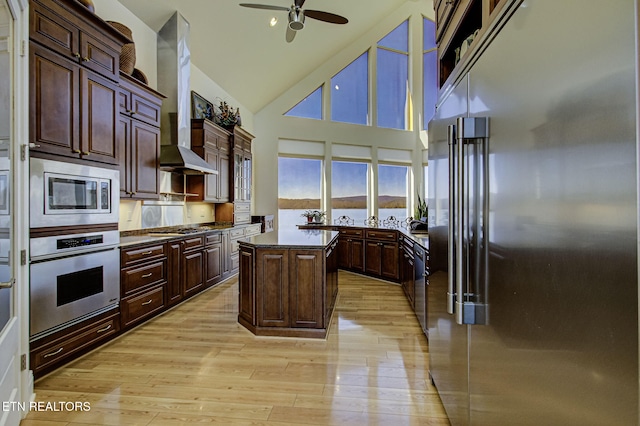 kitchen with a ceiling fan, dark countertops, light wood-style flooring, appliances with stainless steel finishes, and wall chimney range hood