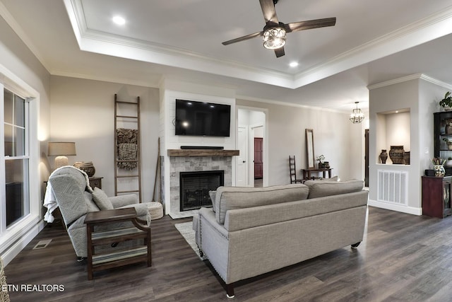 living room with a stone fireplace, dark wood-type flooring, ornamental molding, and a raised ceiling