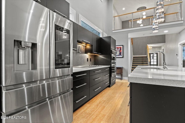 kitchen with appliances with stainless steel finishes, a towering ceiling, sink, hanging light fixtures, and light hardwood / wood-style flooring