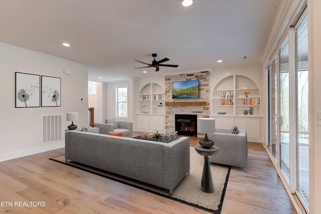 living room with crown molding, built in features, ceiling fan, a stone fireplace, and light wood-type flooring