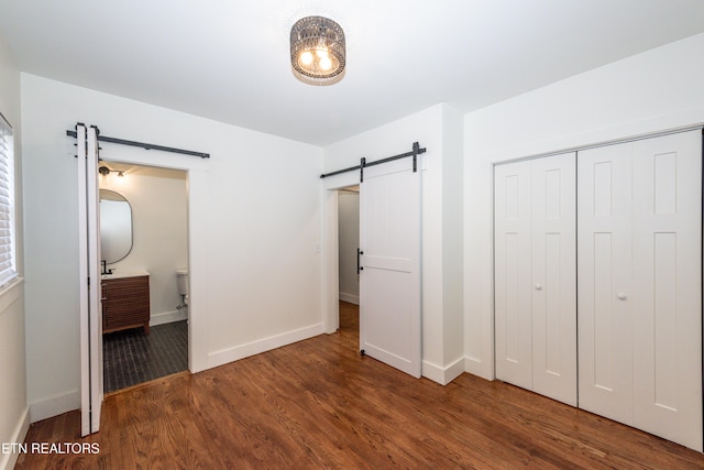 unfurnished bedroom featuring a barn door, baseboards, connected bathroom, dark wood-type flooring, and a closet