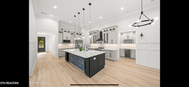 kitchen featuring appliances with stainless steel finishes, wine cooler, hanging light fixtures, a kitchen island with sink, and wall chimney range hood