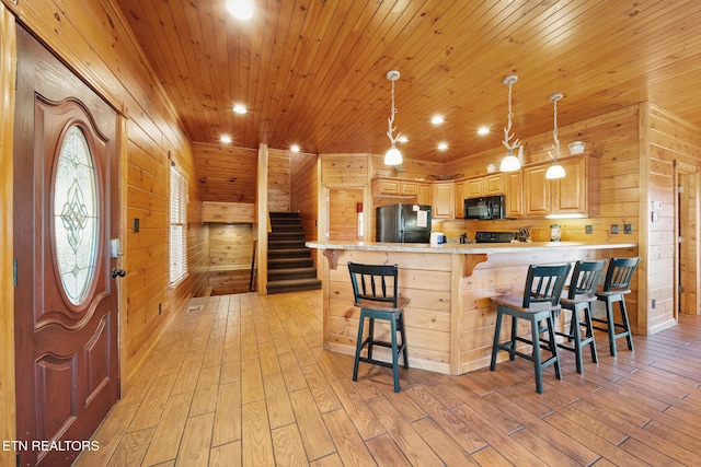 kitchen featuring light brown cabinetry, a breakfast bar area, wood walls, hanging light fixtures, and black appliances