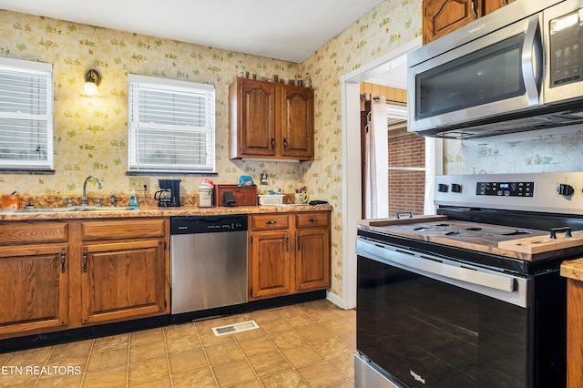 kitchen featuring brown cabinets, light countertops, appliances with stainless steel finishes, a sink, and wallpapered walls