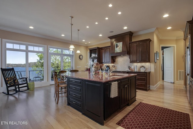 kitchen with sink, light stone counters, decorative light fixtures, an island with sink, and custom range hood