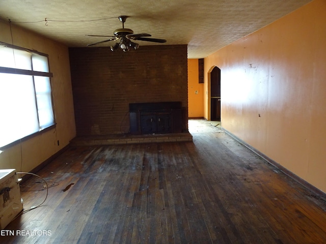 unfurnished living room featuring arched walkways, a textured ceiling, dark wood-style flooring, a ceiling fan, and a brick fireplace