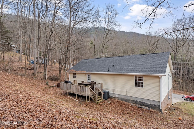 rear view of house featuring central air condition unit, a shingled roof, a deck, and a view of trees