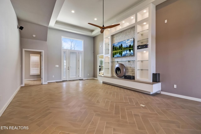 unfurnished living room featuring ceiling fan, a high ceiling, a tray ceiling, built in shelves, and parquet flooring