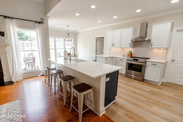 kitchen featuring an island with sink, sink, white cabinets, stainless steel range with electric cooktop, and wall chimney range hood