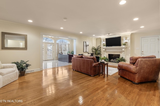 living area with a fireplace with raised hearth, light wood-style flooring, crown molding, french doors, and recessed lighting