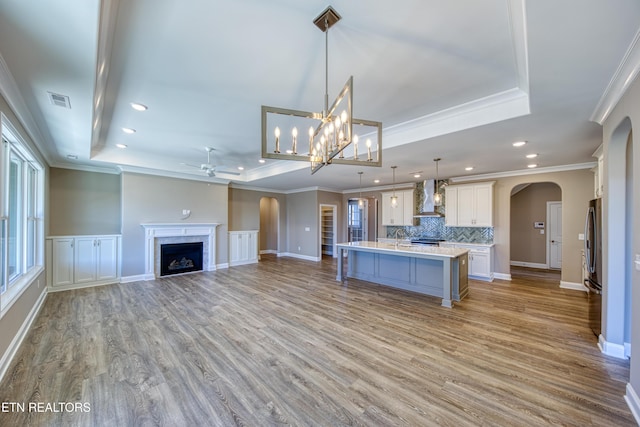 kitchen featuring pendant lighting, a tray ceiling, wall chimney exhaust hood, and a center island with sink