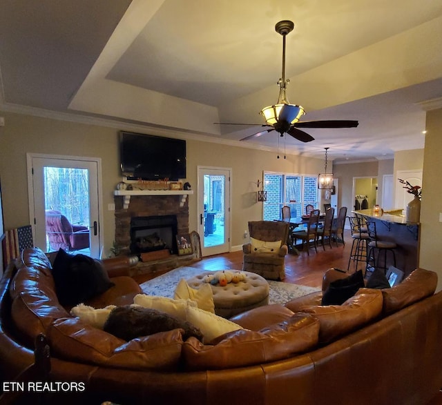 living room with a stone fireplace, crown molding, a tray ceiling, hardwood / wood-style flooring, and ceiling fan