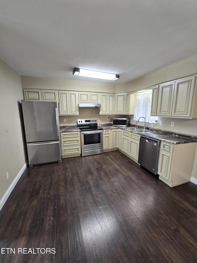 kitchen with cream cabinets, under cabinet range hood, baseboards, appliances with stainless steel finishes, and dark wood finished floors