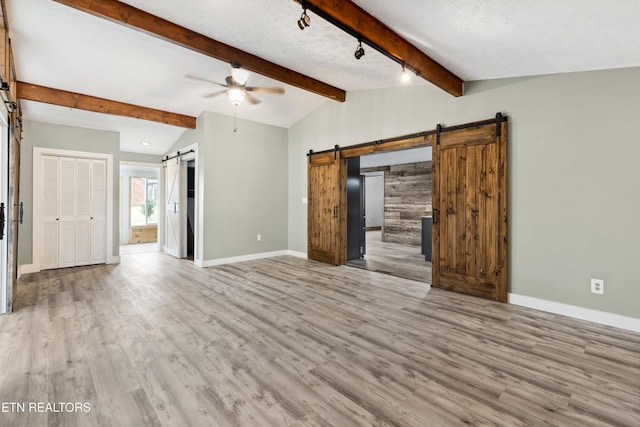 unfurnished living room featuring ceiling fan, a barn door, lofted ceiling with beams, and light wood-type flooring