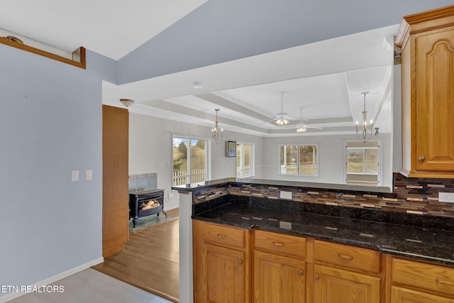 kitchen with dark stone countertops, tasteful backsplash, ceiling fan with notable chandelier, a raised ceiling, and a wood stove