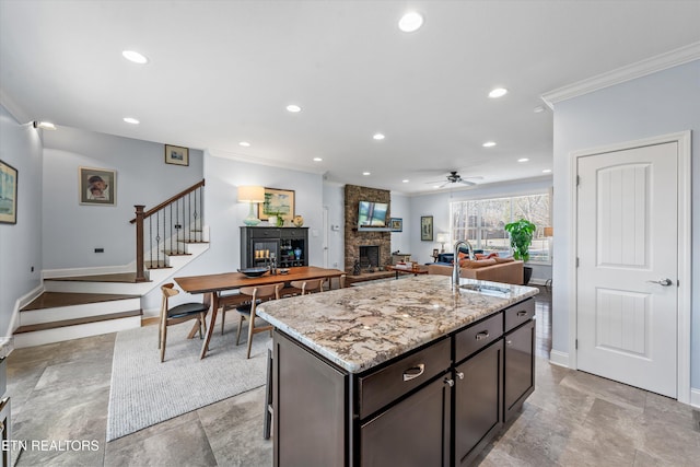 kitchen with dark brown cabinets, ornamental molding, a fireplace, light stone countertops, and a kitchen island with sink