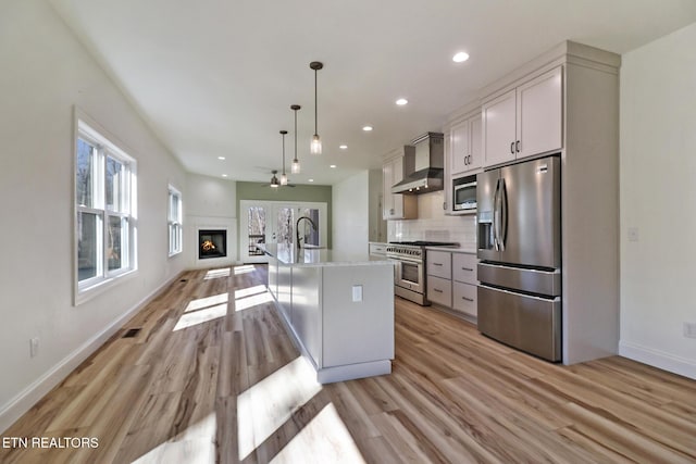 kitchen featuring pendant lighting, wall chimney range hood, appliances with stainless steel finishes, a kitchen island with sink, and backsplash