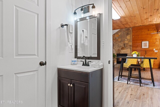 bathroom featuring wood walls, a stone fireplace, wooden ceiling, wood-type flooring, and vanity