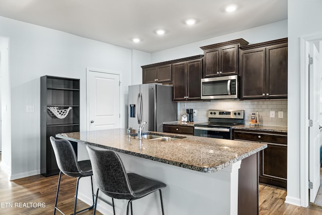 kitchen with sink, appliances with stainless steel finishes, dark brown cabinets, light stone counters, and an island with sink