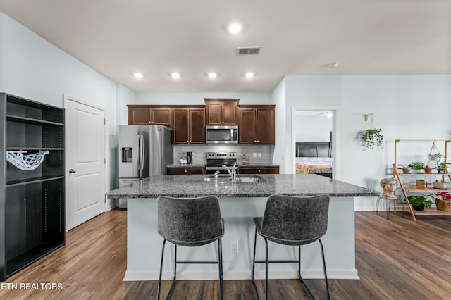 kitchen featuring tasteful backsplash, dark stone countertops, stainless steel appliances, dark wood-type flooring, and a center island with sink