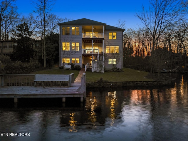 back house at dusk featuring a deck with water view, a balcony, and a yard