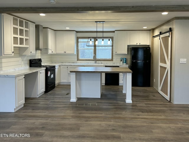 kitchen with wall chimney range hood, hanging light fixtures, black appliances, white cabinets, and a barn door