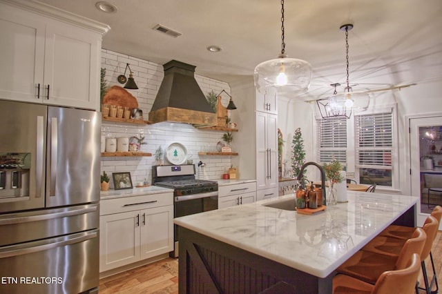 kitchen featuring appliances with stainless steel finishes, hanging light fixtures, custom range hood, white cabinetry, and a kitchen island with sink