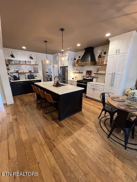 kitchen featuring appliances with stainless steel finishes, custom exhaust hood, light hardwood / wood-style floors, white cabinets, and an island with sink