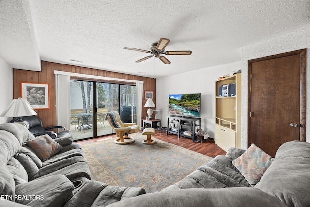 living room with dark wood-type flooring, ceiling fan, wooden walls, and a textured ceiling