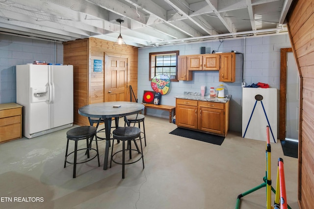 kitchen featuring concrete floors, hanging light fixtures, brown cabinets, white fridge with ice dispenser, and concrete block wall