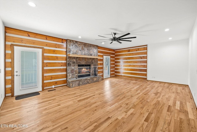 unfurnished living room featuring wooden walls, ceiling fan, a stone fireplace, and light wood-type flooring