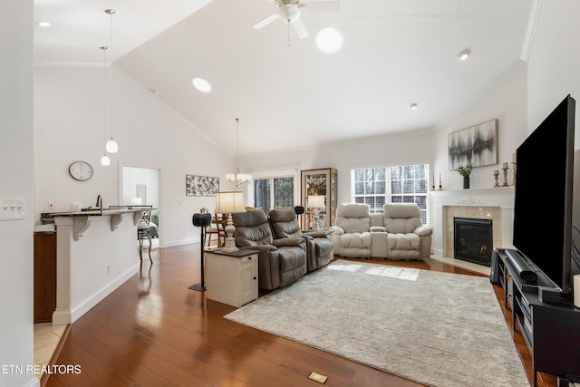 living room with ceiling fan with notable chandelier, hardwood / wood-style floors, high vaulted ceiling, a tiled fireplace, and ornamental molding