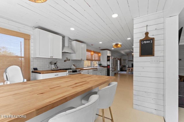 kitchen with stainless steel appliances, white cabinetry, wall chimney range hood, and wood counters