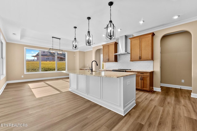 kitchen featuring sink, hanging light fixtures, light wood-type flooring, a kitchen island with sink, and wall chimney range hood