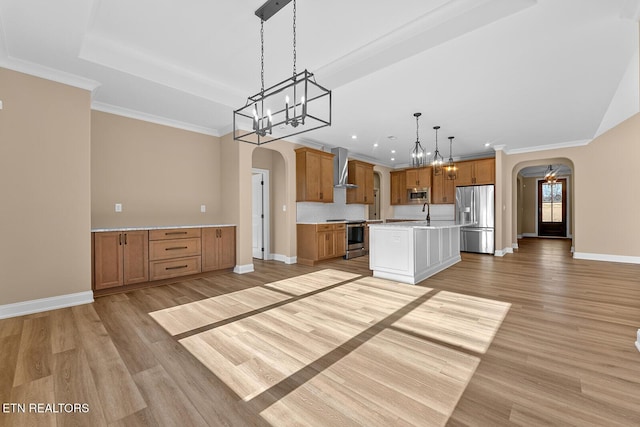 kitchen with wall chimney range hood, hanging light fixtures, stainless steel appliances, light hardwood / wood-style floors, and an island with sink