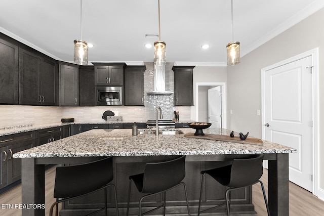 kitchen featuring an island with sink, stainless steel microwave, crown molding, pendant lighting, and a sink