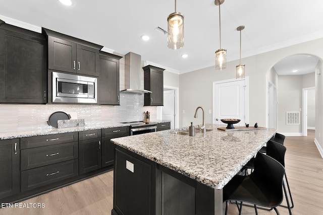 kitchen featuring a kitchen island with sink, a sink, a kitchen breakfast bar, appliances with stainless steel finishes, and wall chimney range hood