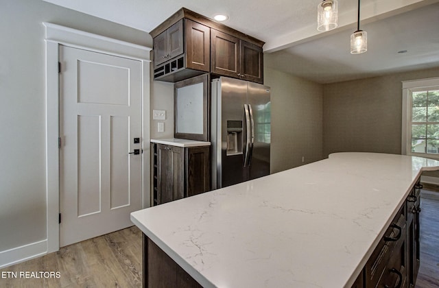 kitchen featuring wood finished floors, hanging light fixtures, dark brown cabinetry, light stone countertops, and stainless steel fridge with ice dispenser