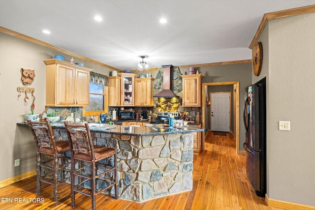 kitchen with crown molding, a breakfast bar area, black appliances, wall chimney exhaust hood, and light wood-type flooring