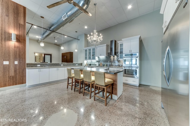 kitchen featuring a towering ceiling, stainless steel appliances, wall chimney exhaust hood, white cabinets, and a large island with sink