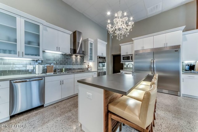 kitchen featuring an island with sink, built in appliances, white cabinets, and wall chimney exhaust hood