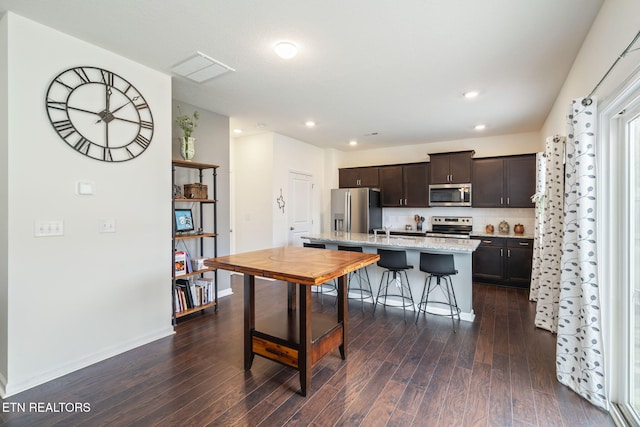 kitchen with dark wood-type flooring, dark brown cabinets, a center island with sink, appliances with stainless steel finishes, and a kitchen breakfast bar